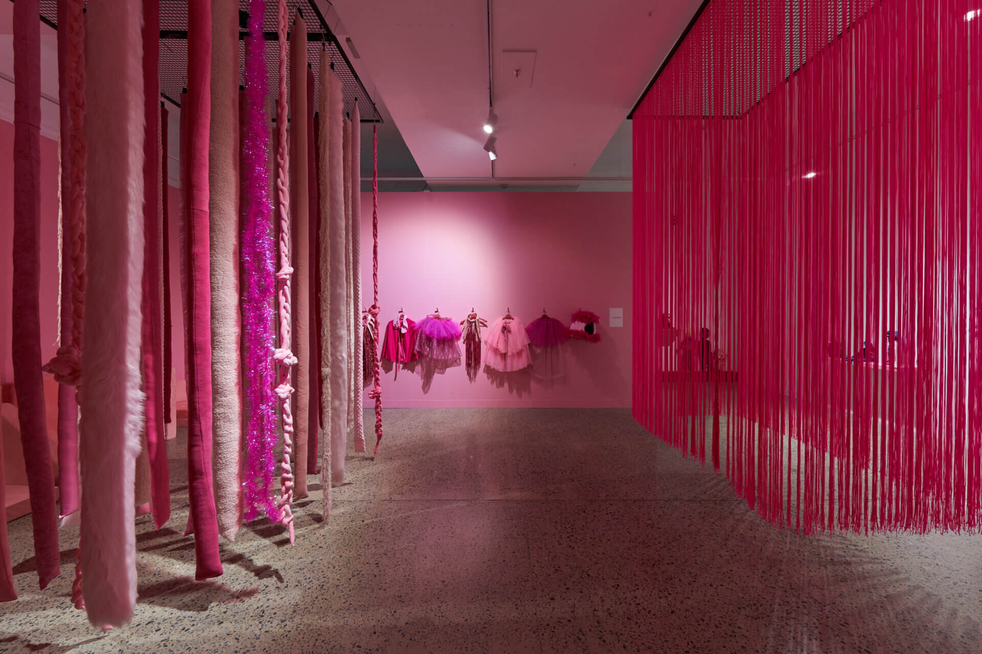 Installation view of a contemporary art exhibition dominated by shades of pink. Long, textured fabric and yarn pieces hang from the ceiling on the left, while a dense curtain of pink cords creates a partition on the right. Pink garments and sculptural textile works are displayed on the back wall in a softly lit gallery with a terrazzo floor.